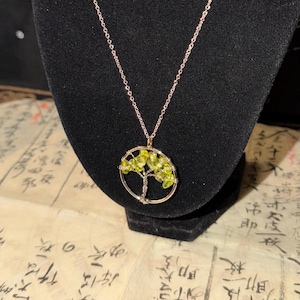 May include: A necklace featuring a circular pendant with a wire-wrapped tree design. The tree branches are adorned with small, translucent lime green gemstone chips. The pendant hangs from a thin, metallic chain, creating a delicate piece of jewelry.