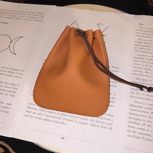 Puede incluir: Una pequeña bolsa de cuero color canela con un cordón marrón oscuro. La bolsa está sobre un libro con texto e ilustraciones. La bolsa es de diseño sencillo y clásico, adecuada para llevar objetos pequeños.