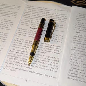 Puede incluir: Un bolígrafo con cuerpo de madera de color rojo oscuro, detalles de metal dorado con un patrón geométrico intrincado y una tapa negra con un clip dorado. El bolígrafo se muestra con la tapa quitada, descansando sobre una página de libro.