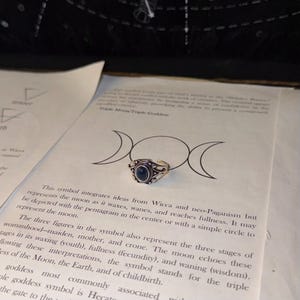 May include: A silver-toned metal ring featuring an oval-shaped dark blue cabochon stone at its center. The ring band has an open design and is decorated with intricate metalwork patterns surrounding the central blue stone.
