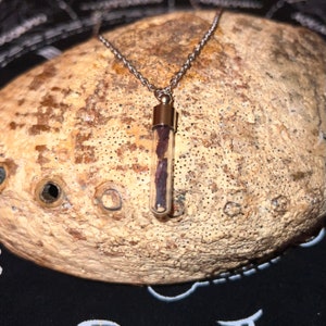 May include: A silver-toned necklace featuring a small, clear glass vial pendant. The vial contains a dark, reddish-brown substance. The pendant is suspended from a delicate chain. The background is a textured, light brown surface.