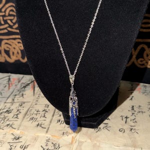 Op de afbeelding: Een zilverkleurige ketting met een puntige, cilindervormige lapis lazuli hanger. De steen is diepblauw met subtiele spikkels en is gevat in een ingewikkelde, zilverkleurige metalen kap in filigraanstijl die naar boven toe taps toeloopt.
