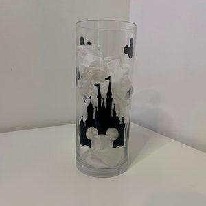May include: A clear glass vase featuring a black silhouette of Cinderella's castle and Mickey Mouse ears. The vase is decorated with black Mickey Mouse ears around the top.