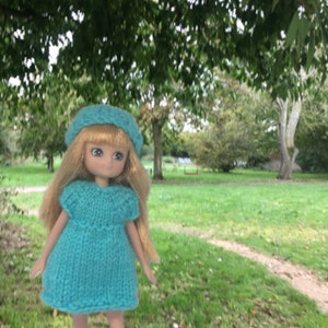 May include: A doll wearing a blue knitted dress and a blue knitted hat. The doll is standing in a grassy area with trees in the background.