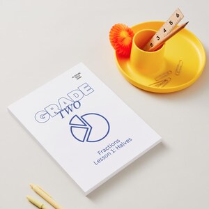 Könnte beinhalten: Ein weißes Arbeitsbuch mit dem Titel "Grade Two" mit einem blauen Bruchteildiagramm und dem Text "Brüche Lektion 1: Hälften". Ein gelber Schreibtisch-Organizer enthält ein Lineal, einen Bleistift und Büroklammern. Eine orangefarbene Papierdekoration befindet sich in der Nähe.