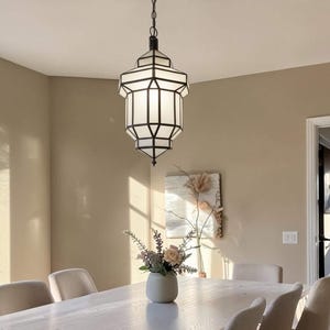 May include: A dining room featuring a light wood table with six beige chairs. A black and white geometric pendant light hangs above the table. A vase of flowers sits in the center of the table. The room has beige walls and a patterned rug.