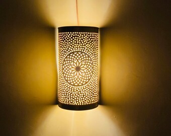 May include: A gold metal wall sconce with a circular cutout design. The sconce is lit and casting a warm glow on the wall.