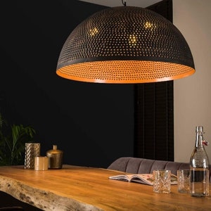 May include: A black metal dome pendant light with a perforated design and a warm orange interior. The light is hanging over a wooden table with glasses and a book on it.