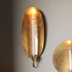 May include: Two gold metal wall sconces with a candle holder in the shape of an oval and a circle. The oval sconce has a white candle inside. The circle sconce has a white candle inside. The sconces are on a white wall.