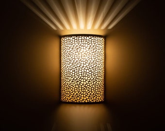 May include: A cylindrical wall sconce with a patterned design, emitting a warm, golden light. The light fixture is mounted on a dark wall, with light rays radiating upwards. The sconce's design creates a unique lighting effect.