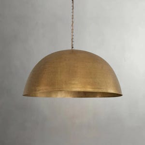 May include: A large, dome-shaped pendant light fixture with a smooth, brushed brass finish. The fixture is suspended from a thin, matching metal chain that extends upward from the center of the dome, creating a clean and uniform appearance.