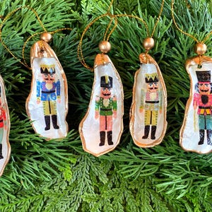 May include: Five Christmas ornaments featuring nutcracker soldier designs. Each ornament is crafted from a natural oyster shell, painted with a gold rim, and adorned with a miniature nutcracker figure in various colors. The ornaments hang from gold cords.