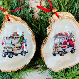 May include: Two Christmas ornaments made from oyster shells. Each shell has a gold-painted edge and a red ribbon for hanging. One ornament features a golf cart loaded with gifts and a Christmas tree. The other shows Santa Claus driving a golf cart filled with presents.