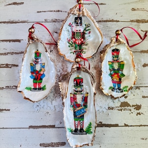 Peut inclure: Quatre coquilles d'huîtres peintes en or avec des rubans rouges et dorés. Chaque coquille présente une illustration vintage différente de casse-noisette.