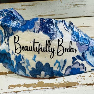 Puede incluir: Objeto decorativo de forma irregular con un estampado floral azul y blanco. El texto "Beautifully Broken" está escrito en el centro en escritura negra. Dos estrellas de mar blancas a cada lado.