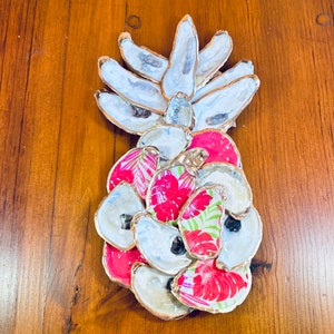 Puede incluir: Una decoración de pared con forma de piña hecha de conchas de ostras. Las conchas están pintadas de blanco y tienen un borde dorado. La piña está decorada con papel estampado floral rosa y verde.