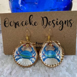 May include: A pair of dangle earrings featuring white scallop shells painted with blue crabs. The shells are edged in gold and hang from gold-toned hooks. The earrings are displayed on a brown card with the text Ocracoke Designs printed in black.