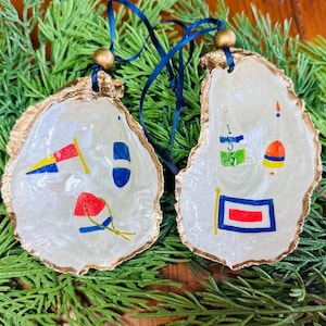 Puede incluir: Dos adornos de concha de ostra con diseños de banderas náuticas. Las conchas tienen bordes pintados de dorado y están ensartadas con cinta azul. Los diseños pintados incluyen banderas, boyas y números en rojo, azul, amarillo y verde.