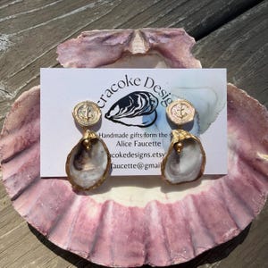 May include: A pair of dangle earrings featuring natural oyster shells with gold-toned metal accents. Each earring has a circular stud top embossed with an anchor design and a small gold bead connecting the shell. The earrings are displayed on a white card.