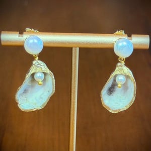 May include: A pair of drop earrings featuring a white spherical pearl stud attached to a gold-toned oyster shell charm. Inside each shell, a smaller white pearl is suspended from a gold-toned post, creating a layered, organic, and metallic aesthetic.