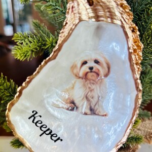 May include: A white oyster shell Christmas ornament with a gold rim. The ornament features a painted image of a small, white dog with the name "Keeper" written below the dog.