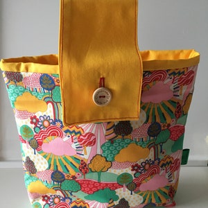 May include: A yellow fabric tote bag with a large pocket on the front. The pocket has a button with the text "Handmade with love". The bag is lined with a yellow fabric and has a colourful floral print with rainbows and clouds.