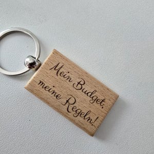 Könnte beinhalten: Holzschlüsselanhänger mit einem silbernen Ring und dem Text "Mein Budget, meine Regeln!" auf der Vorderseite.