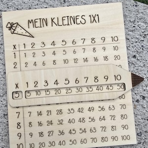 Könnte beinhalten: Multiplikationstabelle aus Holz mit dem Text "MEIN KLEINES 1X1" und einem stiftförmigen Schieber. Die Tabelle zeigt die Multiplikationsreihen von 1 bis 10, mit Fokus auf die 5er-Reihe. Ein lustiges Lernwerkzeug.