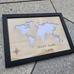 Könnte beinhalten: Ein gerahmter Weltkarte aus Holz mit schwarzem Rahmen. Die Karte ist ausgeschnitten und zeigt einen weißen Hintergrund. Die Namen "Patrick & Angelika" und das Datum "01.10.2023" sind auf das Holz gedruckt. Eine Windrose befindet sich in der linken unteren Ecke.