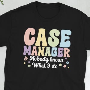 Könnte beinhalten: Schwarzes T-Shirt mit den Worten "CASE MANAGER" in Pastellfarben und dem Satz "Nobody knows What I do" darunter. Das Design enthält florale Akzente. Eine Kamera, eine Brille, ein Telefon und eine Brieftasche befinden sich im Hintergrund.