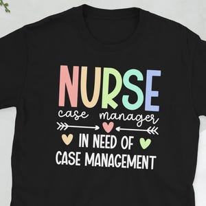 Könnte beinhalten: Schwarzes T-Shirt mit dem Text "NURSE case manager IN NEED OF CASE MANAGEMENT" in Pastellfarben. Das Design beinhaltet Pfeile und Herzsymbole. Eine Kamera, eine Brille, ein Telefon und eine Brieftasche befinden sich im Hintergrund.