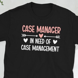 Könnte beinhalten: Schwarzes T-Shirt mit dem Text "CASE MANAGER" in Rosa, darüber "IN NEED OF CASE MANAGEMENT" in Weiß. Das Design beinhaltet rosa Herzen und Pfeile. Ein lässiges T-Shirt für Case Manager.