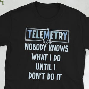 May include: Black t-shirt with the text "Telemetry tech - Nobody knows what I do until I don't do it" in white and light blue. The shirt is short-sleeved and has a crew neck. A camera, glasses, a wallet, and a phone are in the background.