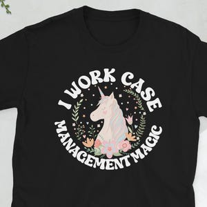 Pode incluir: Camiseta preta com um design circular com um unicórnio e o texto "I WORK CASE MANAGEMENT MAGIC". O unicórnio é branco e rosa, rodeado por flores e folhas. O texto é branco.