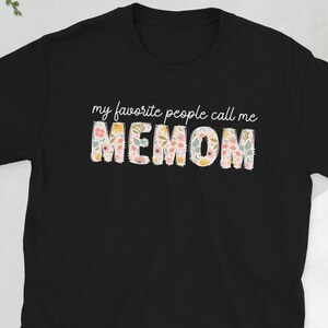 Könnte beinhalten: Schwarzes T-Shirt mit dem Text "my favorite people call me MEMOM" in weißer Schrift über dem Wort "MEMOM". Das Wort "MEMOM" ist in einem floralen Design mit rosa, gelben und grünen Blumen.