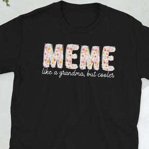 Op de afbeelding: Zwart T-shirt met het woord "MEME" in een bloemenpatroon. De tekst "like a grandma, but cooler" staat onder het woord "MEME". Het shirt is gemaakt van een zacht materiaal en heeft korte mouwen.