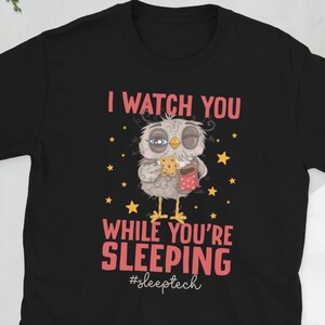 Peut inclure: T-shirt noir avec un dessin de hibou de dessin animé. Le hibou tient un biscuit et une tasse, avec le texte "I WATCH YOU WHILE YOU'RE SLEEPING #sleeptech". Le design est rose et blanc avec des étoiles.