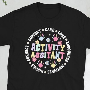 May include: Black t-shirt featuring the words "Activity Assistant" in a colourful design, encircled by words such as "Support" and "Love." The design incorporates handprints and floral elements.