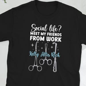 Puede incluir: Camiseta negra con el texto "Social life? Meet my friends from work" en blanco, junto con gráficos de instrumentos médicos y el nombre "Kelly Allis Rich" en azul claro. Una camiseta informal.
