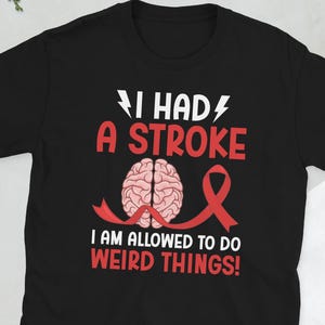 May include: Black t-shirt featuring the text "I HAD A STROKE" in white and red, a brain graphic, and a red ribbon. The phrase "I AM ALLOWED TO DO WEIRD THINGS!" is printed below. A comfortable, everyday tee.