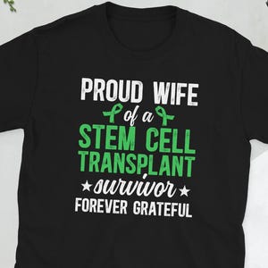 May include: Black t-shirt with white and green text. The text reads "PROUD WIFE of a STEM CELL TRANSPLANT survivor FOREVER GRATEFUL". The shirt has a crew neck and short sleeves.