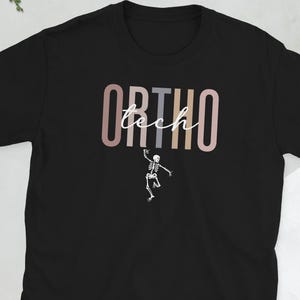Könnte beinhalten: Schwarzes T-Shirt mit dem Schriftzug "ORTHO tech" in einem Farbverlauf und einer kleinen weißen Skelettgrafik darunter. Das kurzärmelige Shirt ist aus weichem Material.