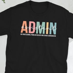 May include: Black t-shirt with the word "ADMIN" in colorful block letters and "assistant" in cursive. Below, the text reads "(n) PROFESSIONAL PROBLEM SOLVER AND CHAOS COORDINATOR". A camera, glasses, and a phone are visible.