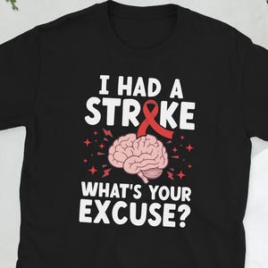 Peut inclure: T-shirt noir avec l'inscription "I HAD A STROKE" au-dessus d'un dessin de cerveau et les mots "WHAT'S YOUR EXCUSE ?" en blanc. Le cerveau est rose et un ruban rouge est inclus dans le design.