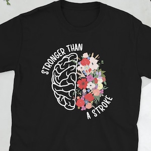 Puede incluir: Camiseta negra con un gráfico de cerebro blanco y un diseño floral. El texto "STRONGER THAN A STROKE" está arqueado alrededor del cerebro y las flores. Una camiseta informal para el uso diario.