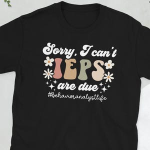 Op de afbeelding: Zwart t-shirt met de tekst "Sorry, I can't, IEPs are due" in retro stijl. Het ontwerp bevat bloemmotieven en de hashtag #behavioranalystlife. Een casual t-shirt voor gedragsanalisten.