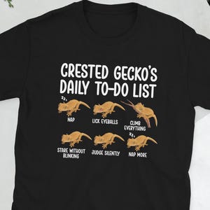 Peut inclure: T-shirt noir avec l'inscription blanche "CRESTED GECKO'S DAILY TO-DO LIST". Le t-shirt présente des illustrations de geckos avec des phrases comme "NAP", "LICK EYEBALLS" et "CLIMB EVERYTHING".
