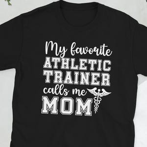 Pode incluir: Camiseta preta com texto branco: "My favorite ATHLETIC TRAINER calls me MOM" e um símbolo médico. A camiseta é de manga curta e feita de um material macio.