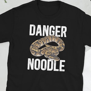 Op de afbeelding: Zwart T-shirt met de witte tekst "DANGER NOODLE" en een gedetailleerde illustratie van een bruine, beige en zwarte slang. De slang is opgerold in het midden van het ontwerp.