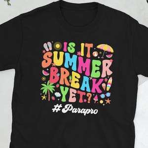 May include: Black t-shirt with a colorful graphic that reads "IS IT SUMMER BREAK YET?" in various fonts and colors. The design includes images of a palm tree, a sun umbrella, and other summer-themed elements. The hashtag #Parapro is below the text.